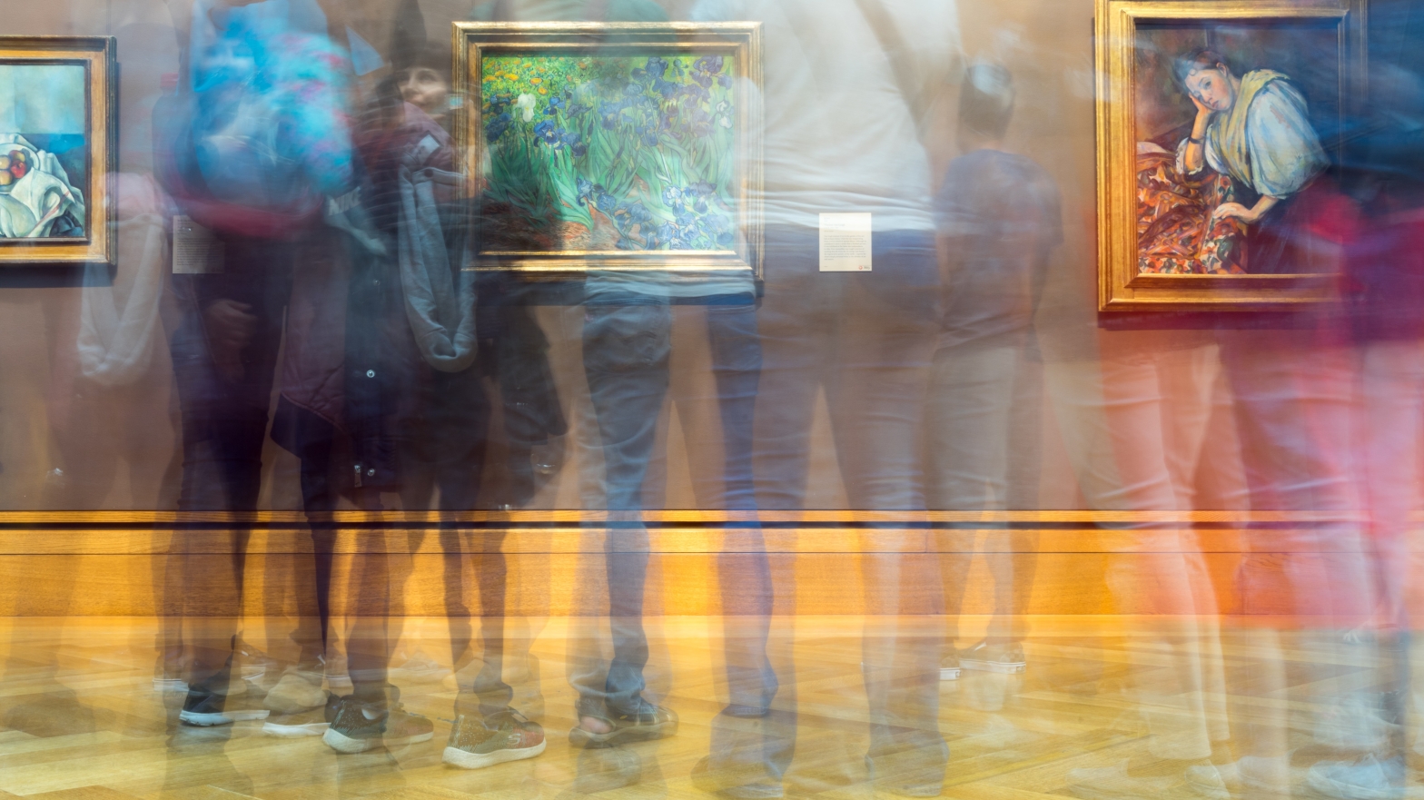 Image shows a group of people standing in front of paintings in an art gallery. The photo has been taken with slow shutter speed so that the people are blurred and the paintings are in focus.